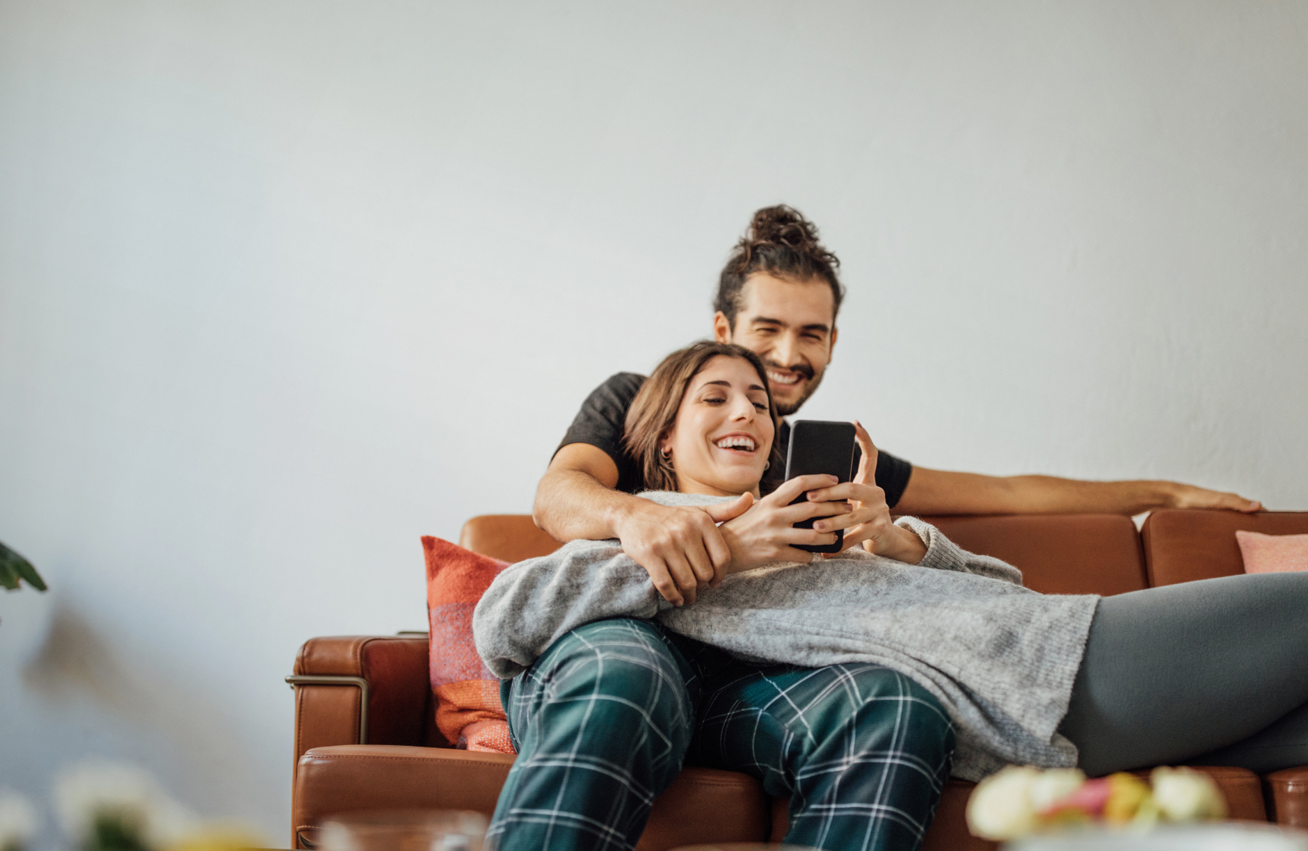 Smiling young woman using smart phone while reclining on boyfriend sitting on sofa at home